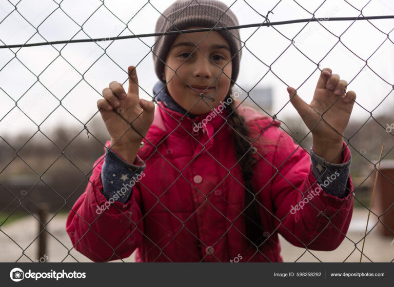 A little girl with a sad look behind a metal fence. Social problem of refugees and forced migrants